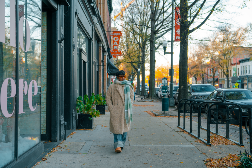 Black woman walking on sidewalk wearing cozy green scarf, and long tan sherpa coat